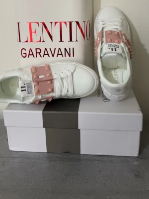 Vita sneakers Valentino Garavani - Stilrena vita sneakers från Valentino Garavani med rosa band och nitar över vristen. Skorna har klassisk låg profil, rund tå och är tillverkade i slätt läder. Guldlogga på innersulan och cool design som sticker ut.