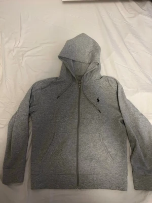 Grå zip hoodie från Polo Ralph Lauren - Snygg grå hoodie från Polo Ralph Lauren med dragkedja framtill, två fickor och klassisk huva. Tillverkad i mjuk bomullsblandning och dekorerad med den ikoniska lilla svarta logon på bröstet. Perfekt för en chill och stilren look.