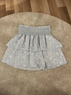 Blå volangkjol från UNIQ Studio - Supersöt kort blå kjol från UNIQ Studio med volanger och smock i midjan. Kjolen har ett ljust paisleymönster och är perfekt för dig som gillar en feminin och trendig stil. Lätt och luftig känsla, passar perfekt till varmare dagar.