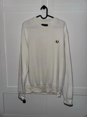 Vit sweatshirt från Fred Perry - Klassisk vit sweatshirt från Fred Perry med rund halsringning och svart broderad logga på bröstet. Tröjan har ribbade muddar med svarta detaljer vid ärmslut och nederkant. Två väldigt små ljusa fläckar framtill, syns om man tittar noga (Bild 3)