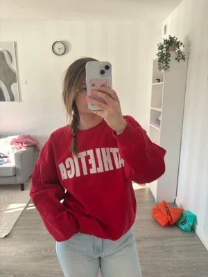 Röd sweatshirt från Gina Tricot - Cool röd sweatshirt från Gina Tricot med vit ATHLETICA-tryck på framsidan. Tröjan har en avslappnad passform, ribbade muddar och är gjord i mjukt material som känns skönt mot huden. Perfekt för en sportig och casual look.