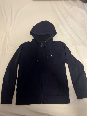 Marinblå hoodie från Polo Ralph Lauren - Snygg marinblå hoodie från Polo Ralph Lauren med dragkedja framtill och klassisk logga broderad på bröstet. Hoodien har huva med dragsko och en stor magficka. Tillverkad i mjuk bomullsblandning som är skön att bära hela dagen.
