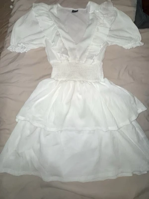 Studentklänning  - Supersöt vit kortklänning från Gina Tricot med volangdetaljer, v-ringning och smock i midjan. Klänningen har korta puffärmar och dubbla lager i kjolen för extra volym. Perfekt för sommarens alla tillfällen!