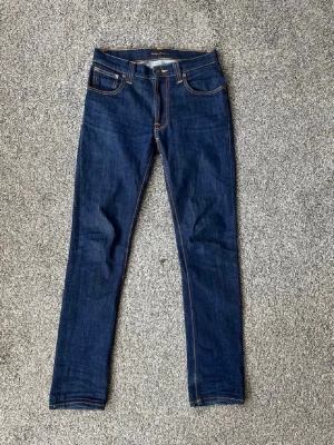 Mörkblå skinny jeans från Nudie - Snygga mörkblå skinny jeans från Nudie Jeans med klassiska kontrastsömmar och femficksmodell. Jeansen har en diskret logga på bakfickan och är tillverkade i mjuk denim med stretch för extra komfort. Perfekta till en avslappnad streetstyle-look.