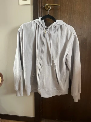 Ljusgrå zip hoodie med huva - En enkel och clean ljusgrå hoodie med dragkedja framtill och stor ficka på magen, från H&M. Hoodien har justerbar huva med snören och ribbade muddar vid ärmar och nederkant. Säljer för att den inte används. Storlek S.
