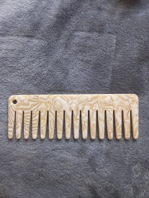 Bredtandad beige kam i plast - Säljer en bredtandad kam i beige med marmorerat mönster. Tillverkad i plast och har ett hål i hörnet för enkel upphängning. Perfekt för att reda ut håret utan att slita. Snygg accessoar till badrumshyllan eller väskan.