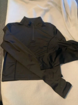 Svart träningströja med halv dragkedja - Säljer en svart långärmad träningströja med hög krage och halv dragkedja framtill. Tillverkad i ett stretchigt funktionsmaterial som passar perfekt för löpning eller gym. Enkel och stilren design utan synligt märke.