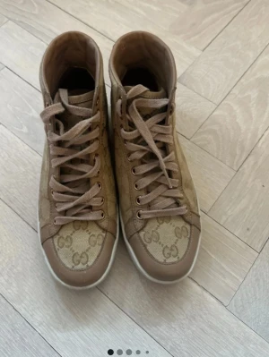 Beige höga sneakers från Gucci - Säljer ett par snygga höga sneakers från Gucci i beige canvas med klassiskt GG-mönster och detaljer i ljusbrunt läder. Skorna har vita sulor, guldfärgade öljetter och matchande beigea skosnören. Perfekta för dig som gillar exklusiv streetstyle.
