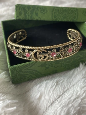 Gucci armband med rosa stenar - Säljer ett lyxigt armband i guld och färgad metall. Armbandet har ett öppet, flätat mönster med små blommor och dekorerat med rosa stenar. Perfekt statement piece för dig som gillar exklusiva accessoarer och detaljer. Helt nytt! Justerbart. Inspirerat. 🌸 FRI FRAKT! Passa på! 