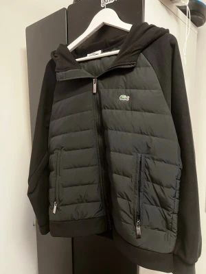 Lacoste Cardigan - Snygg svart pufferjacka från Lacoste med huva och dragkedja framtill. Jackan har quiltad front och rygg, mjuka ärmar i sweatshirtmaterial och två dragkedjeförsedda fickor. Liten grön Lacoste-logga på bröstet ger en clean look.