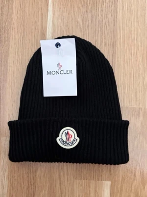 Svart ribbstickad mössa Moncler - Säljer en svart ribbstickad mössa från Moncler med uppvikt kant och klassisk Moncler-logga framtill. Mössan är tillverkad i mjukt material och har en stilren, enkel design som passar perfekt till höst och vinter.
