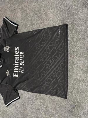 Real Madrid matchtröja - Snygg svart Real Madrid matchtröja från Adidas med vita detaljer och diskret mönster över hela tyget. Tröjan har klubbmärke och Adidas-logga på bröstet samt 'Emirates Fly Better' tryckt framtill. Perfekt för fotbollsträning eller att bära till vardags.