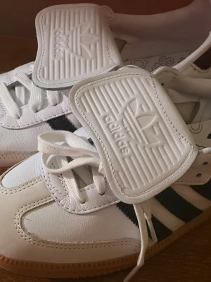 Adidas vita sneakers med svarta ränder - Snygga vita Adidas sneakers med klassiska svarta ränder på sidorna och gummisula. Skorna har en unik stor vit övervikande plös med Adidas-logga i relief och vita skosnören. Är inte använda. Box medföljer ej. 