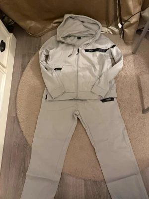 Ljusgrått loungewear-set från BOSS - Snyggt ljusgrått loungewear-set från BOSS med dragkedja på tröjan, huva och svarta detaljer med logga. Setet består av en hoodie och matchande byxor i mjukt material, perfekt för chill dagar hemma.