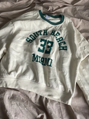 Vit sweatshirt South Beach Miami - Vit sweatshirt med gröna detaljer och tryck på bröstet där det står 'South Beach 33 Miami' i college-stil. Tröjan har rund halsringning med grön ribbad kant och är gjord i mjukt bomullsmaterial. Perfekt för en avslappnad och sportig look.