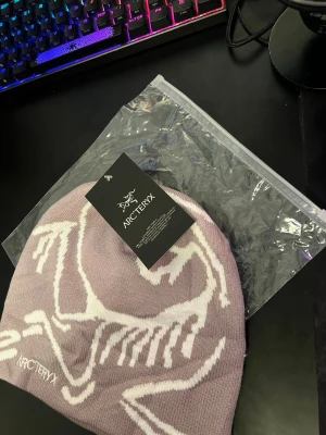 Grej mössa från Arc'teryx - Snygg ljuslila mössa från Arc'teryx med vit logga och grafiskt mönster på framsidan. Tillverkad i mjukt, stickat material som sitter skönt på huvudet. Perfekt för dig som vill ha en stilren och modern accessoar till höst och vinter.