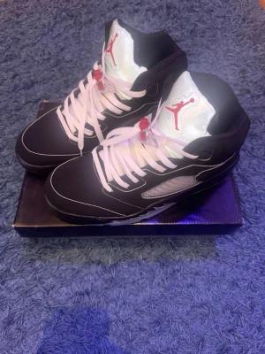 Air Jordan 5 Retro svart/vit - Säljer ett par Air Jordan 5 Retro sneakers i svart mocka med vita och grå detaljer. Skorna har vita skosnören, genomskinliga meshpaneler på sidorna och en ikonisk röd Jumpman-logga på plösen. Sulan är svart och vit med synlig Air-enhet. Perfekta för dig som gillar streetwear.