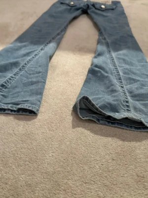 True Religion ljusblå jeans med fickdetaljer - Säljer ett par ljusblå jeans från True Religion med snygga bakfickor och stora silverfärgade knappar. Jeansen har klassisk femficksdesign och tydliga sömmar på fickorna. Perfekta för dig som gillar street och vill sticka ut.