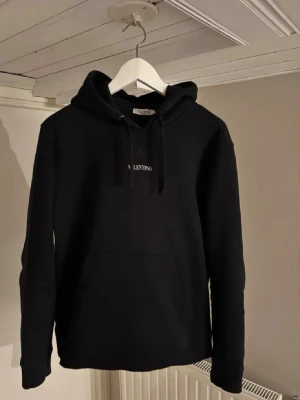 Svart hoodie från Valentino -  Varan är självklart äkta!!Snygg svart hoodie från Valentino med klassisk huva och dragsnören. Diskret logga broderad i vitt på bröstet. Hoodien har en stor magficka och är tillverkad i mjukt bomullsmaterial som känns skönt mot huden. Perfekt för en avslappnad och trendig look.