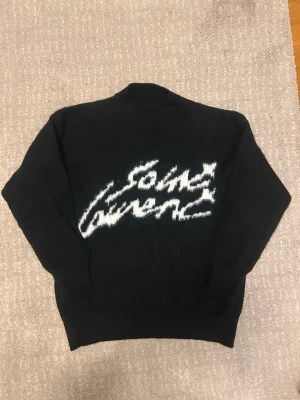 Svart stickad tröja Saint Laurent - Cool svart stickad tröja från Saint Laurent med vit fuzzy logga framtill. Tröjan har rund halsringning och långa ärmar. Perfekt för dig som gillar streetstyle och vill ha något unikt i garderoben.