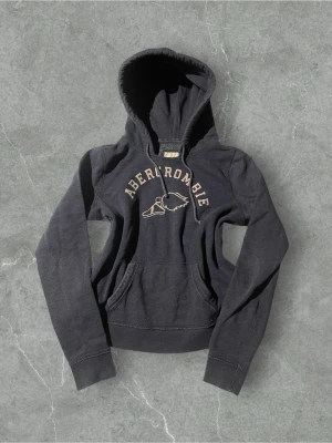 Mörkblå hoodie från Abercrombie | 40/L - Snygg mörkblå hoodie från Abercrombie med klassisk logga och vinge på bröstet. Hoodien har huva med dragsko och en stor magficka framtill. Tillverkad i mjukt bomullsmaterial som känns skönt mot huden. Perfekt för en avslappnad och sportig stil.