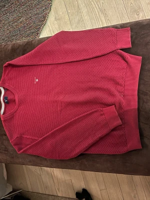 Röd stickad tröja från GANT - Snygg röd stickad tröja från GANT med diskret logga på bröstet. Tröjan har ett strukturerat mönster, rund halsringning och ribbade muddar vid ärmslut och nederkant. Perfekt för dig som vill ha en klassisk men ändå modern look.