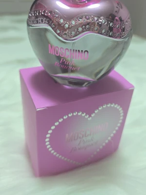 Moschino Pink Bouquet parfym - Moschino Pink Bouquet parfym i en hjärtformad flaska med silver- och rosa detaljer samt glittrande dekorationer. Flaskan står på en rosa kartong med pärlformad hjärtdesign. Perfekt för dig som gillar söta och lekfulla dofter.