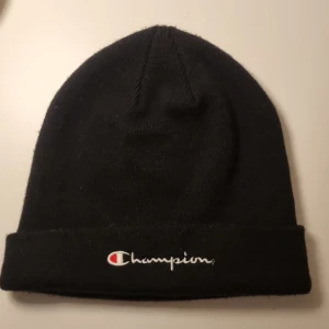 Svart mössa från Champion - Säljer en svart mössa från Champion med broderad logga framtill i vitt och rött. Klassisk modell med uppvikt kant, perfekt för kalla dagar. Enkel och stilren design som passar till det mesta.
