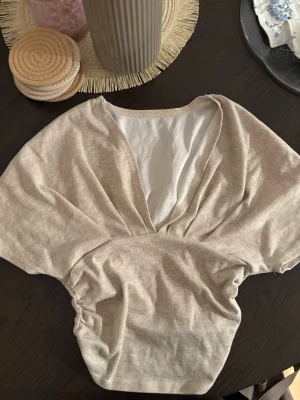 Beige omlottopp med v-ringning - Beige topp med omlott-design och djup v-ringning. Toppen har korta ärmar och är lätt rynkad i midjan. 
