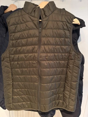 Olivgrön quiltad dunväst - Snygg olivgrön dunväst med quiltat mönster och dragkedja framtill. Västen har hög krage och är lätt vadderad för extra värme. Perfekt att slänga över en hoodie för en casual look. Materialet är troligtvis polyester med dunfyllning.