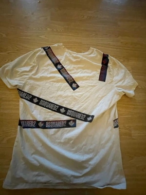 Vit DSQUARED2 t-shirt med banddetaljer - Säljer en vit t-shirt från DSQUARED2 med svarta banddetaljer och logga samt lönnlövsmotiv. T-shirten har korta ärmar och en klassisk rund halsringning. Perfekt för dig som vill sticka ut med en unik design.