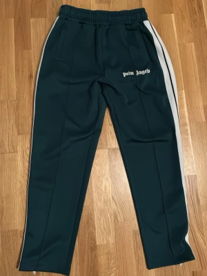 Palm Angels gröna mjukisbyxor - Mörkgröna mjukisbyxor från Palm Angels med vita sidoränder och logga på vänster lår. Byxorna har resår i midjan, raka ben och dragkedjor vid bensluten. Tillverkade i ett glansigt material som ger en sportig vibe.