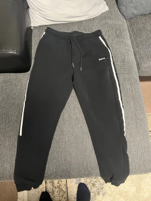 Svarta mjukisbyxor från BOSS - Säljer ett par svarta mjukisbyxor från BOSS med vita ränder längs sidorna och dragsko i midjan. Byxorna har resår vid bensluten och en liten BOSS-logga på vänster sida. Perfekta för chill dagar eller när du vill ha en sportig look.
