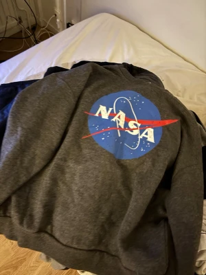Mörkgrå NASA hoodie - Säljer en mörkgrå hoodie med stort NASA-tryck på bröstet. Hoodien har klassisk huva och magficka, samt ribbade muddar vid ärmslut och nederkant. Materialet är mjukt och bekvämt.
