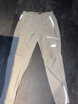 Gråa The North Face byxor. - Säljer ett par beige cargobyxor med elastisk midja och flera fickor, inklusive en klassisk cargoficka på sidan. Byxorna har snygga reflexdetaljer på benen och dragkedja vid benslut för extra stilpoäng. Perfekta för en avslappnad och trendig look.