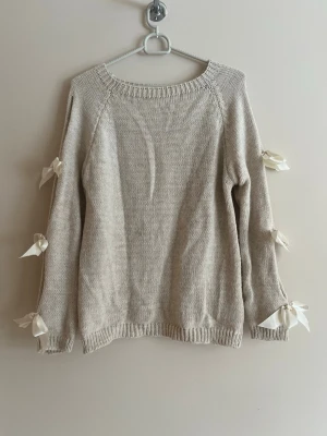 Beige stickad tröja med rosetter - Supersöt beige stickad tröja med rund halsringning och dekorativa vita rosetter längs ärmarna. Tröjan har en avslappnad passform och ribbade muddar vid ärmslut och nederkant. Perfekt för dig som gillar en gullig och trendig stil.