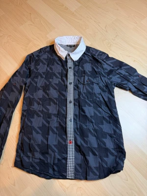 Mönstrad skjorta från KappAhl - Snygg långärmad skjorta från KappAhl med mörkgrått och svart geometriskt mönster, vit krage och rutiga detaljer längs knappslån. Skjortan har bröstficka med knapp och kontrastfärgade knappar för en cool look.