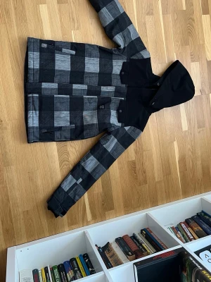 Svart och gråmönstrad vindjacka med luva - Snygg vindjacka med rutigt mönster i svart och grått. Jackan har huva, dragkedja och tryckknappar framtill samt två fickor. Axelpartiet och huvan är helsvarta vilket ger en cool kontrast. Perfekt för dig som gillar streetwear och vill sticka ut. Storlek 164 och den är använd två gånger syns inte på den alls.
