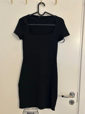 Svart ribbad kortklänning från NLY One - En svart ribbad kortklänning med fyrkantig ringning och korta ärmar från NLY Trend i storlek XS. Klänningen har en slim passform och är tillverkad i ett stretchigt material som sitter snyggt på kroppen. Endast använd en gång. 