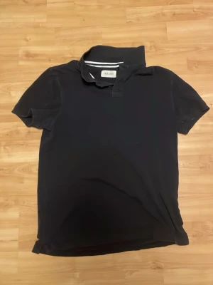 Svart pikétröja från Pier One - Klassisk svart pikétröja från Pier One med korta ärmar och krage. Tröjan har en enkel design utan mönster och är tillverkad i mjukt bomullsmaterial. Perfekt till jeans eller shorts för en clean look.