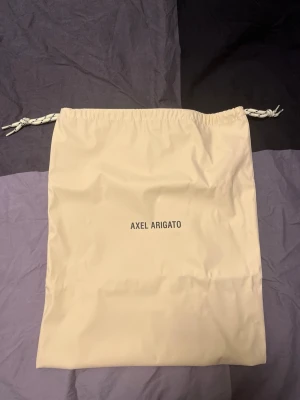 Beige dustbag från Axel Arigato - Säljer en beige dustbag från Axel Arigato med dragsko upptill. Perfekt för att förvara skor eller accessoarer. Enkel design med logga i svart text på framsidan. Tillverkad i mjukt tygmaterial.