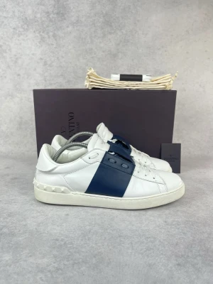Valentino vita sneakers med blå detalj - Snygga vita sneakers från Valentino med bred mörkblå läderdetalj över sidan och klassiska nitar på hälen. Skorna har en låg profil och är tillverkade i slätt läder med vita snören och diskret logga på plösen.