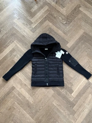 Svart pufferjacka från Moncler - Snygg svart pufferjacka från Moncler med quiltad kropp och stickade ärmar. Jackan har huva, dragkedja framtill och fickor med dragkedja. Moncler-logga på ärmen och extra ficka med dragkedja på ena ärmen. Perfekt för kalla vinterdagar. Passar runt 175-180