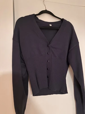 Marinblå ribbad kofta med knappar - Snygg mörkblå kofta med v-ringning och ribbad nederdel. Koftan har långa ärmar och knäppning framtill med svarta knappar. 