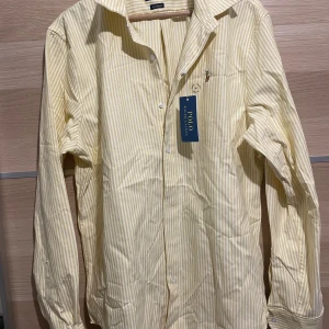 Gul- och vitrandig skjorta Polo Ralph Lauren - Klassisk långärmad skjorta från Polo Ralph Lauren i gul- och vitrandigt bomullstyg. Skjortan har knappar framtill, krage och den ikoniska broderade loggan på bröstet. Perfekt för en stilren och avslappnad look.