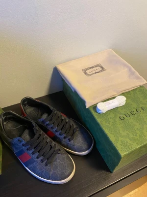 Gucci ace skor - Snygga svarta Gucci sneakers med klassiskt GG-mönster i canvas och läderdetaljer. Skorna har svarta snören och den ikoniska röd-gröna randen på sidan. Tecken på användning men fortfarande i väldigt fint skick. Skriv vid minsta lilla fundering🙌