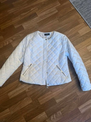 Vit jacka från Flash Woman City - Snygg vit jacka från Flash Woman City med rund halsringning och dragkedja framtill. Jackan har två fickor med dragkedja och tryckknappar vid halsen. Perfekt för dig som gillar stilrena och moderna plagg.