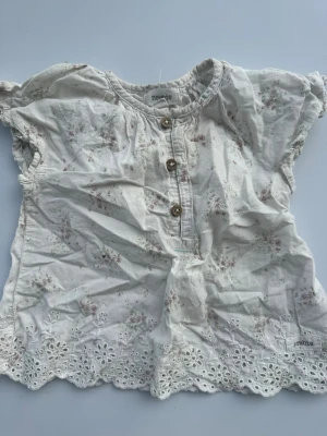 Vit blommig blus från Newbie - Söt vit blus från Newbie med små blommor i rosa och grönt. Blusen har korta puffärmar, träknappar framtill och broderad spetskant längst ner. Tillverkad i mjuk bomull, perfekt för en romantisk och avslappnad stil.fläck finns