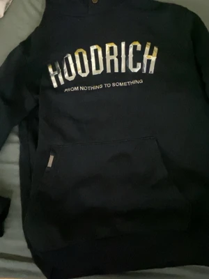 Svart HOODRICH hoodie med tryck - Säljer en svart hoodie från Hoodrich med stor logga i vitt och guld på bröstet och texten 'FROM NOTHING TO SOMETHING' under. Klassisk känguruficka framtill och huva. Perfekt för dig som gillar streetwear och vill ha en snygg och bekväm hoodie.