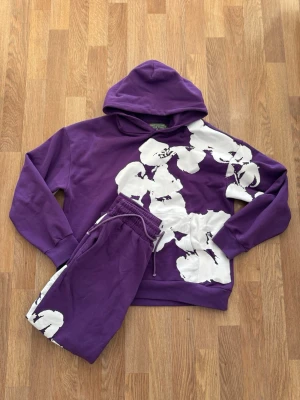 Lila hoodie från Denim Tears med blommor - Säljer en lila hoodie  med stora vita blommor tryckta över framsidan. Hoodien har en klassisk huva och ribbade muddar. Materialet är mjukt och skönt, perfekt för en chill och trendig look. 